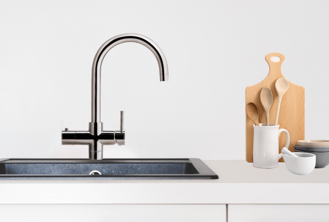 A modern kitchen sink with a stainless steel faucet next to a white countertop holding a cutting board, wooden utensils in a ceramic holder, and stacked bowls.