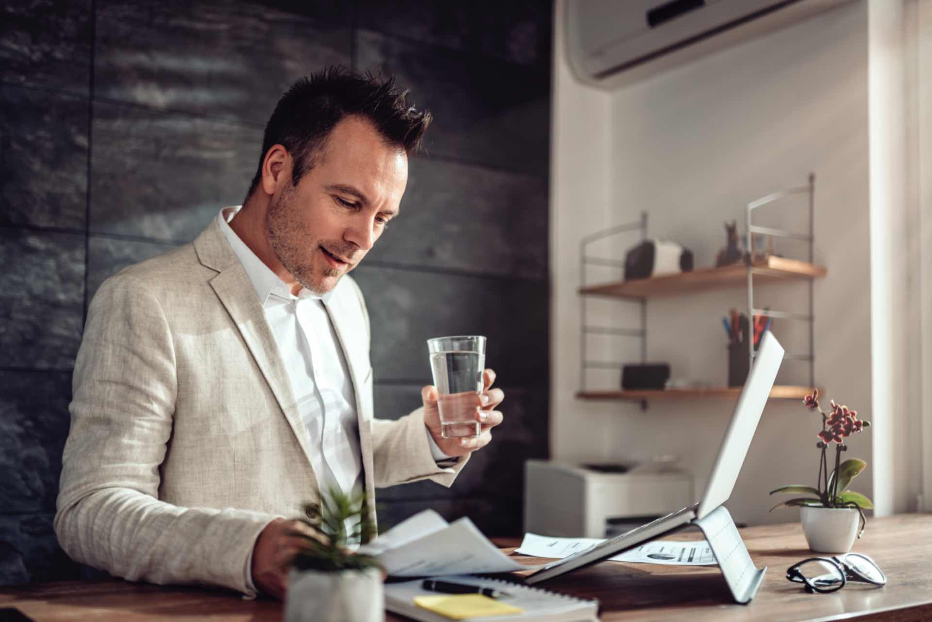 Man in a light blazer holds a glass of water while looking at documents and a laptop on a wooden desk in a modern office setting.
