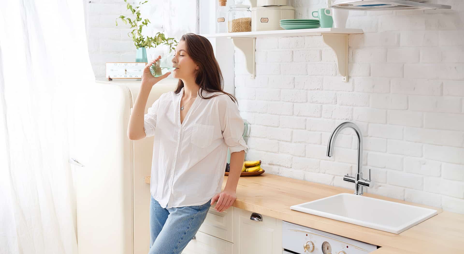 A woman in a white shirt and jeans drinks from a glass while leaning against a kitchen counter in a bright kitchen.