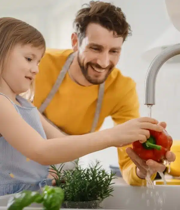 A man and a little girl washing vegetables in a kitchen sink.